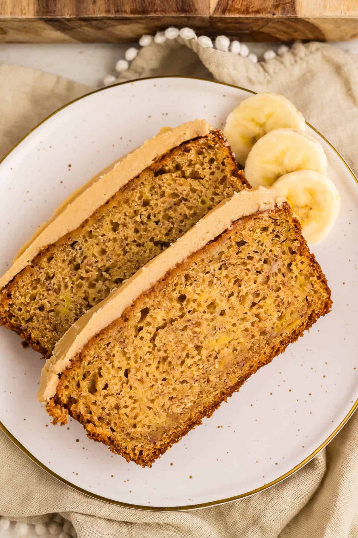 Two slices of banana bread with caramel frosting on a speckled plate, garnished with 3 banana slices on the side.