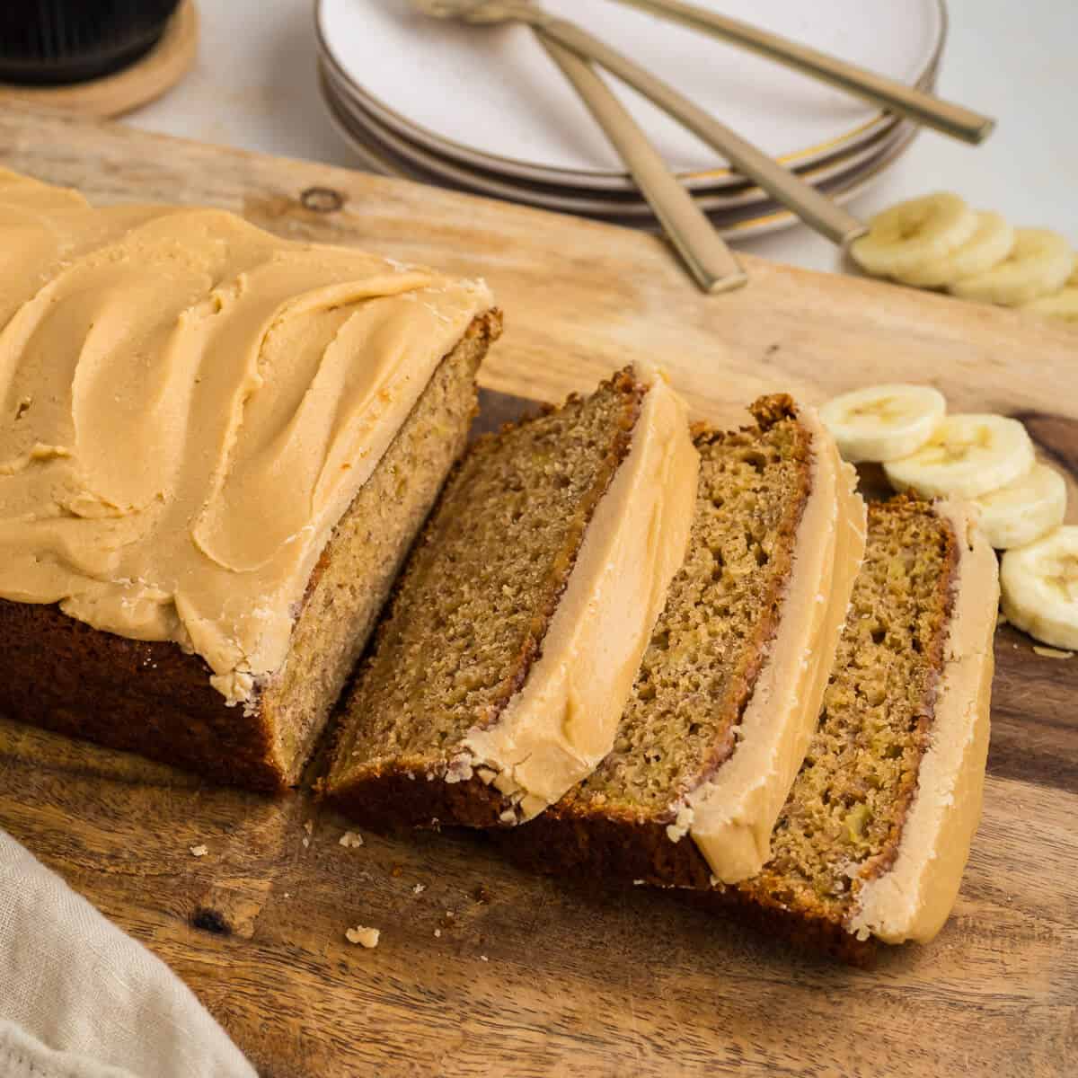 Loaf of banana bread with caramel frosting on top. Three slices have just been cut off the loaf and are laying slightly stacked. There are sliced bananas i the background on the board. There are plates in the background as well.