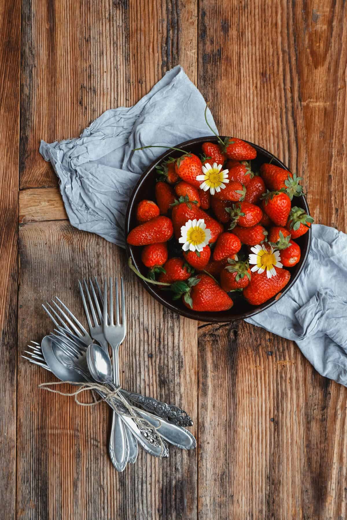Bowl for fresh strawberries with a few small flowers on top on a rustic wood table, some antique flatware next to it tied up in twine.