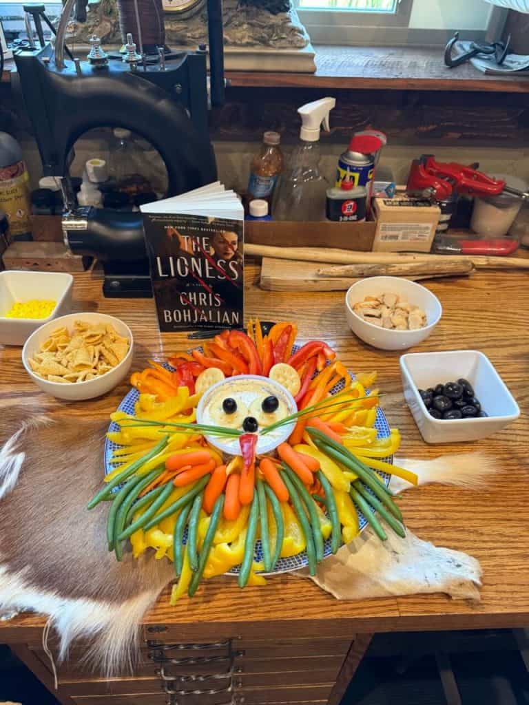 A veggie platter with dip made to look like a lion, a bowl of bugles, animal crackers and chocolate covered raisins. The platter sits on an animal hide, and the book "The Lioness" is behind the platter.