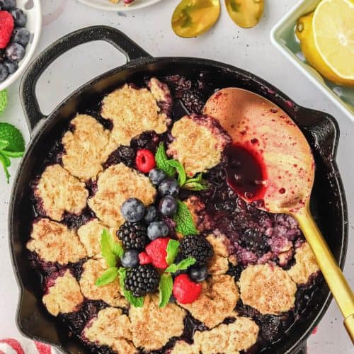 Mixed Berry Cobbler in a cast iron skillet with a scoop missing and a gold serving spoon laying on the side. The cobbler is garnished with fresh berries and mint leaves. There is plated cobbler, gold spoons, berries and lemons surrounding the skillet.