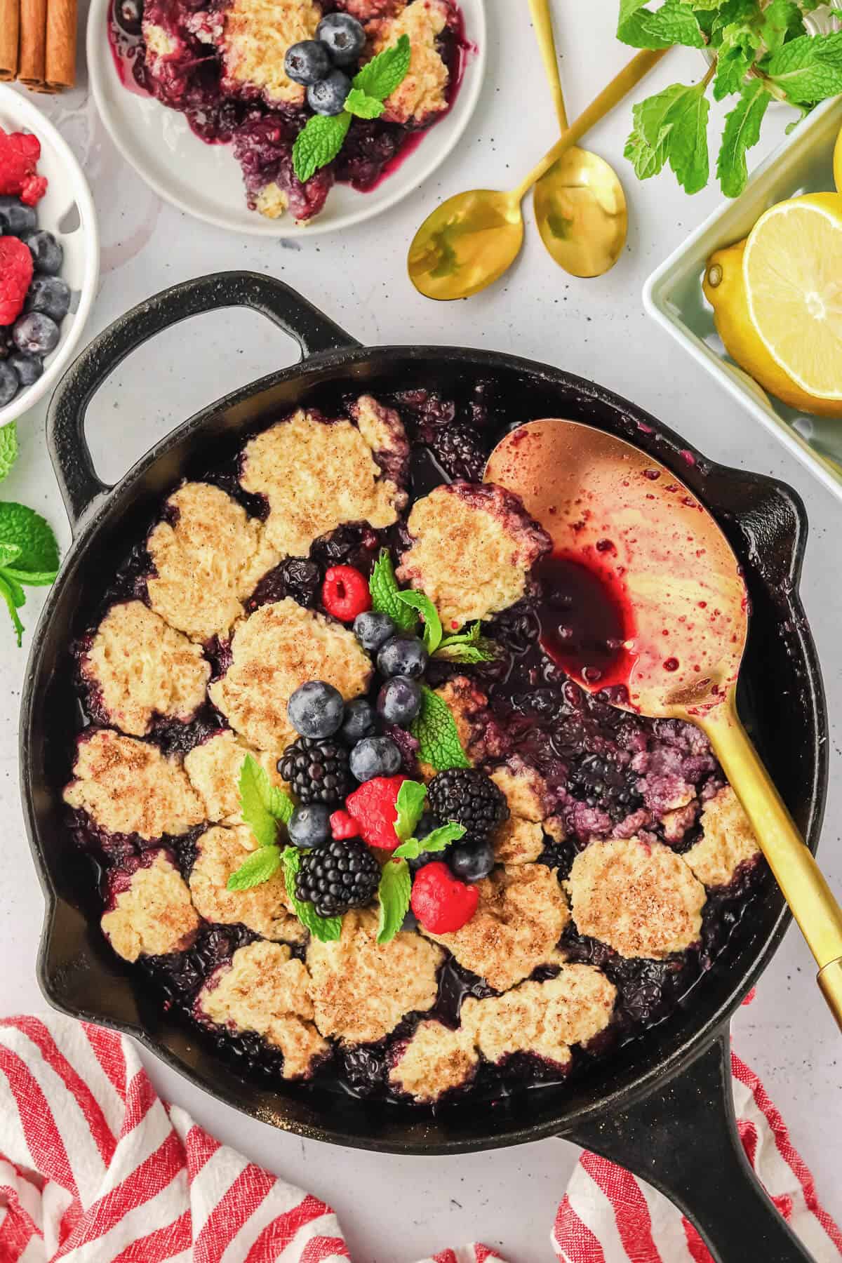 Mixed Berry Cobbler in a cast iron skillet with a scoop missing and a gold serving spoon laying on the side. The cobbler is garnished with fresh berries and mint leaves. There is plated cobbler, gold spoons, berries and lemons surrounding the skillet.