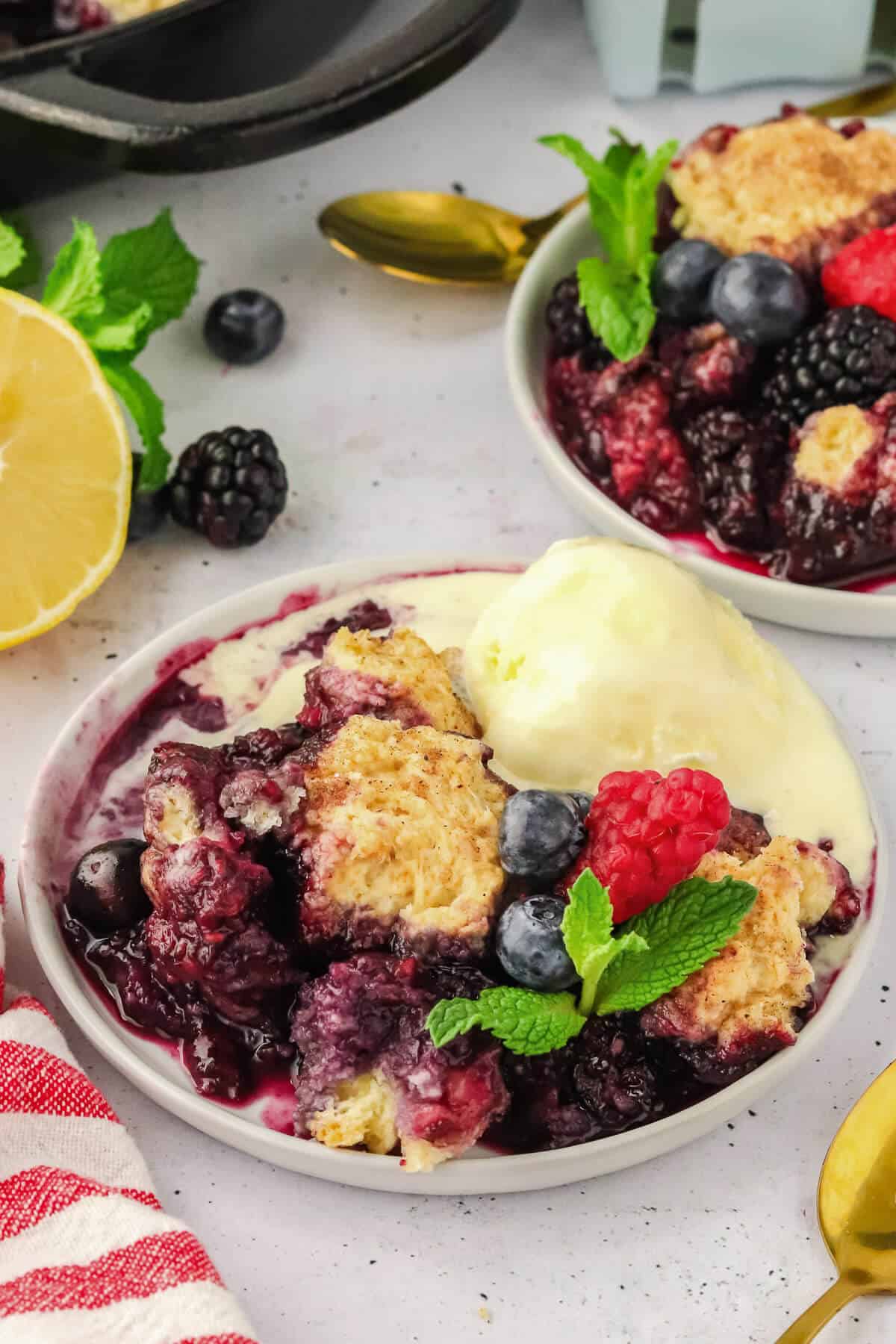 Mixed Berry Cobbler on a white plate with a scoop of ice cream that is beginning to melt. It's garnished with fresh blueberries, raspberries, and mint leaves. There is another plate of cobbler in the background.