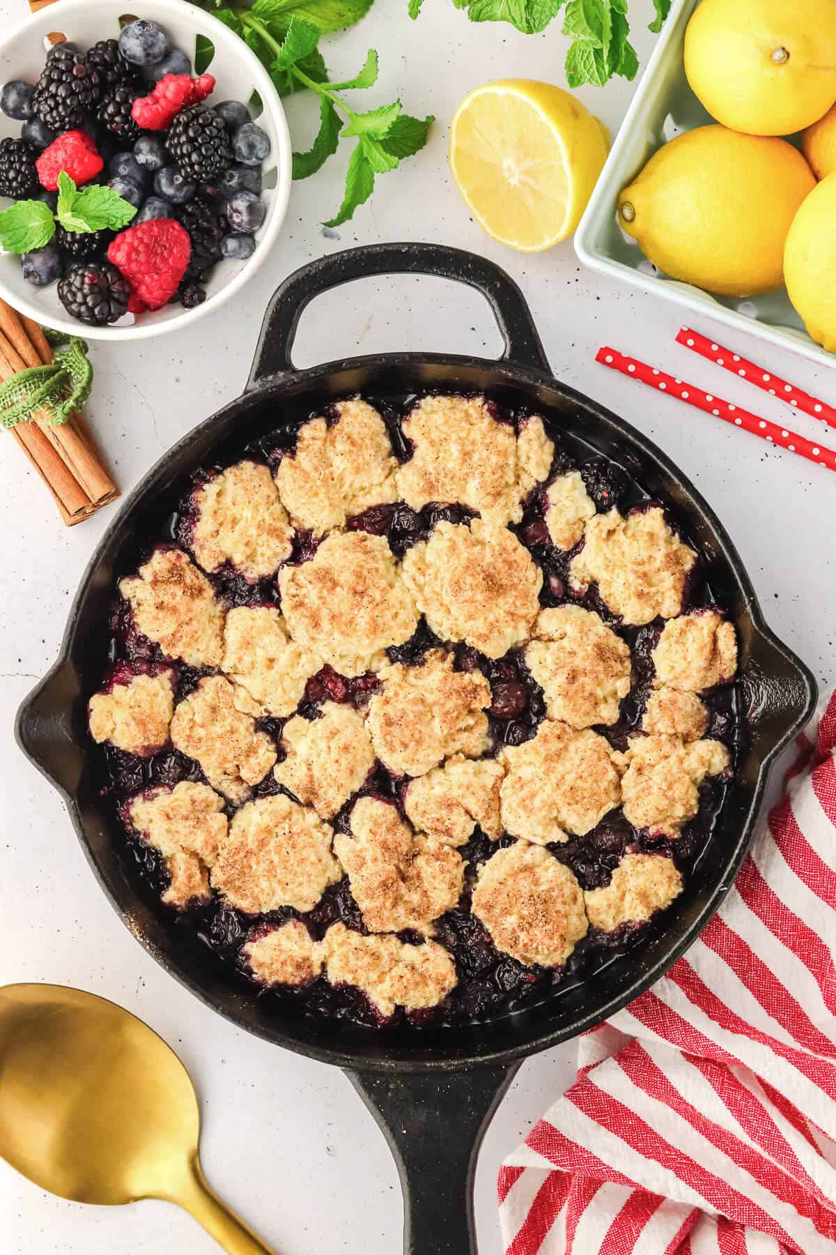 Mixed Berry Cobbler in a cast iron skillet fresh out of the oven. A bowl of fresh berries sits off to the side as well as a group of lemons.