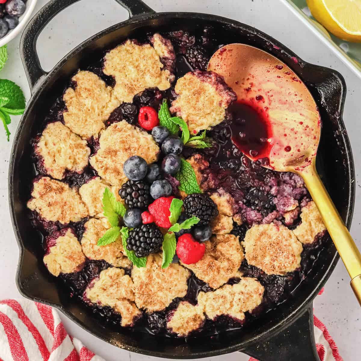 Mixed Berry Cobbler in a cast iron skillet with a scoop missing and a gold serving spoon laying on the side. The cobbler is garnished with fresh berries and mint leaves. There is plated cobbler, gold spoons, berries and lemons surrounding the skillet.