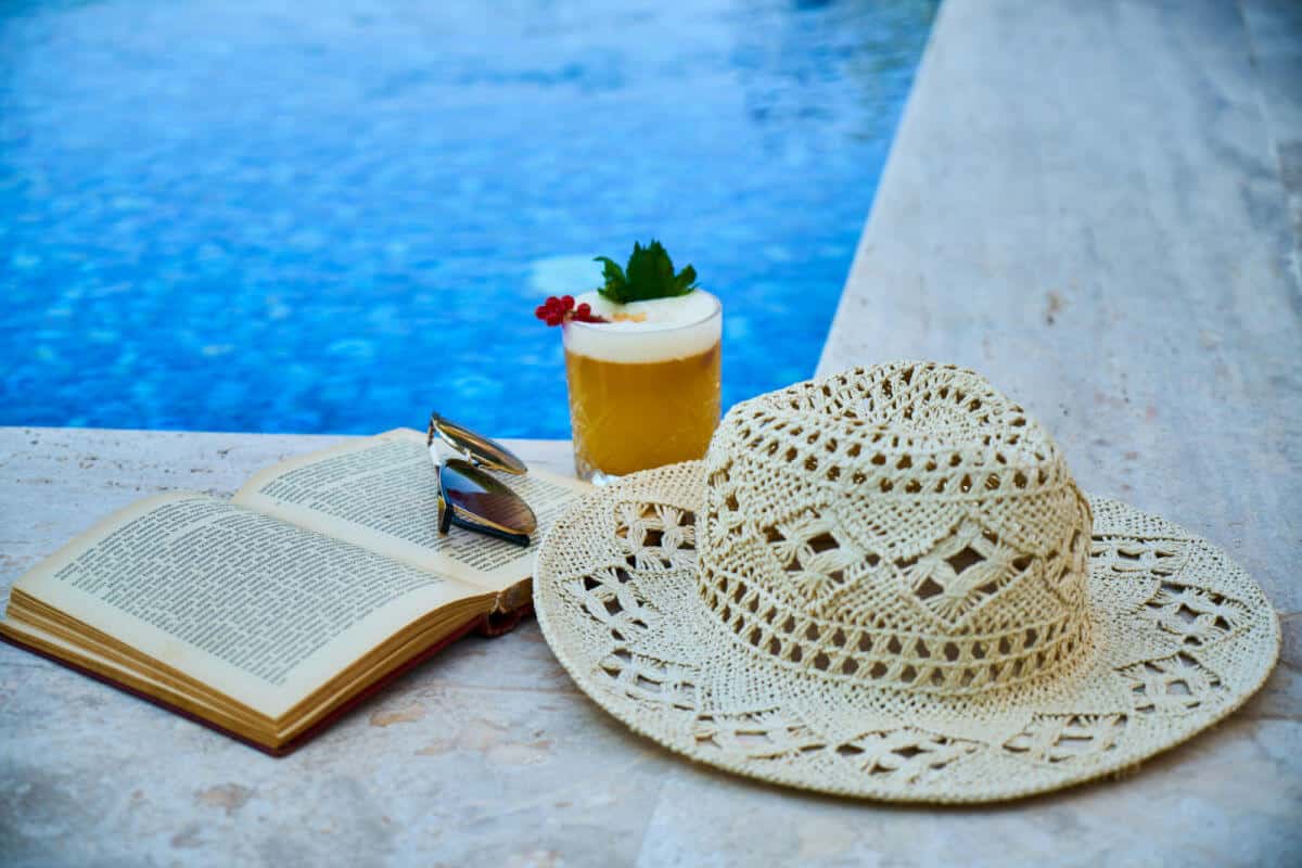Sun hat, open book, sunglasses and a mocktail sitting on the edge of a pool.