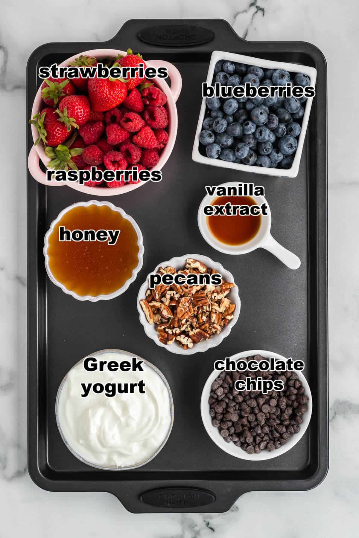 Ingredients to make Yogurt Bark with berries. The ingredients are separated into bowls on a sheetpan. They include: strawberries, raspberries, blueberries, pecans, chocolate chip, Greek yogurt, honey, and vanilla extract.