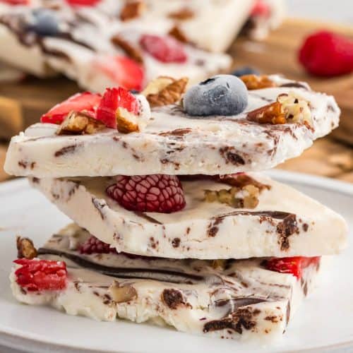 Three squares of Frozen Yogurt Bark stacked on a plate. Remaining Yogurt Bark is in the back ground. The frozen bark has berries, pecans and chocolate in it.