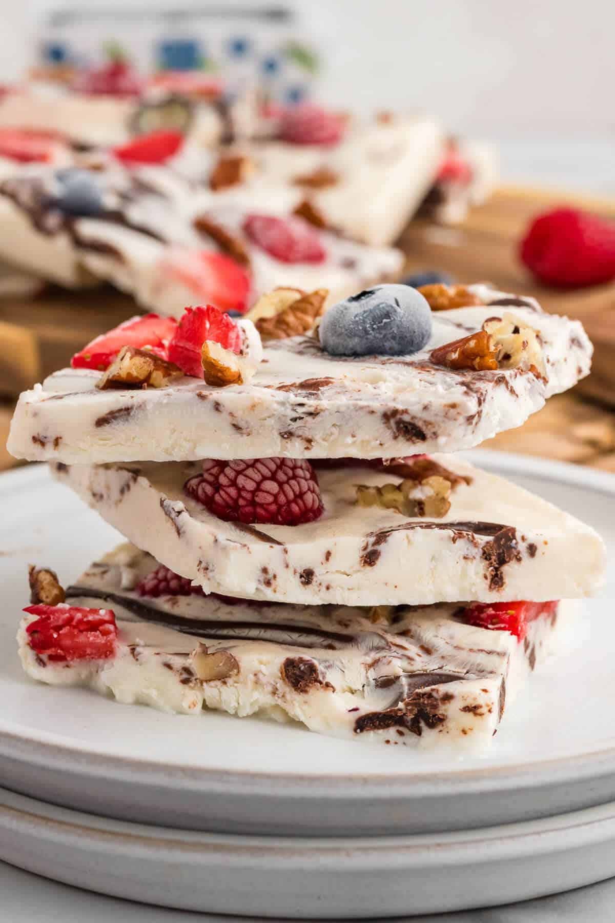 Three squares of Frozen Yogurt Bark stacked on a plate. Remaining Yogurt Bark is in the back ground. The frozen bark has berries, pecans and chocolate in it.