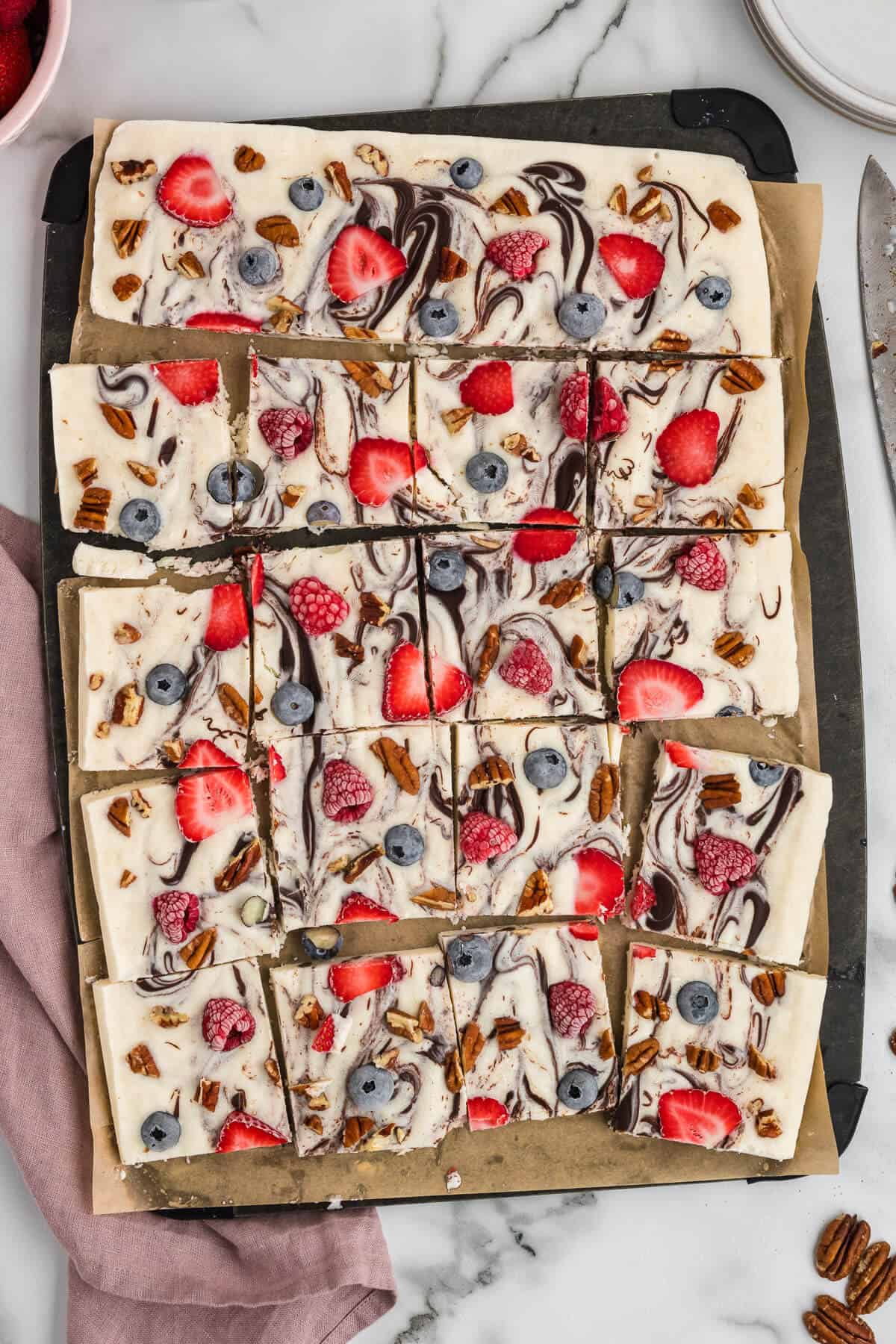 Squares of Frozen Yogurt Bark just cut on a cutting board. The bark has chocolate swirled through and contains strawberries, raspberries, blueberries and pecans.