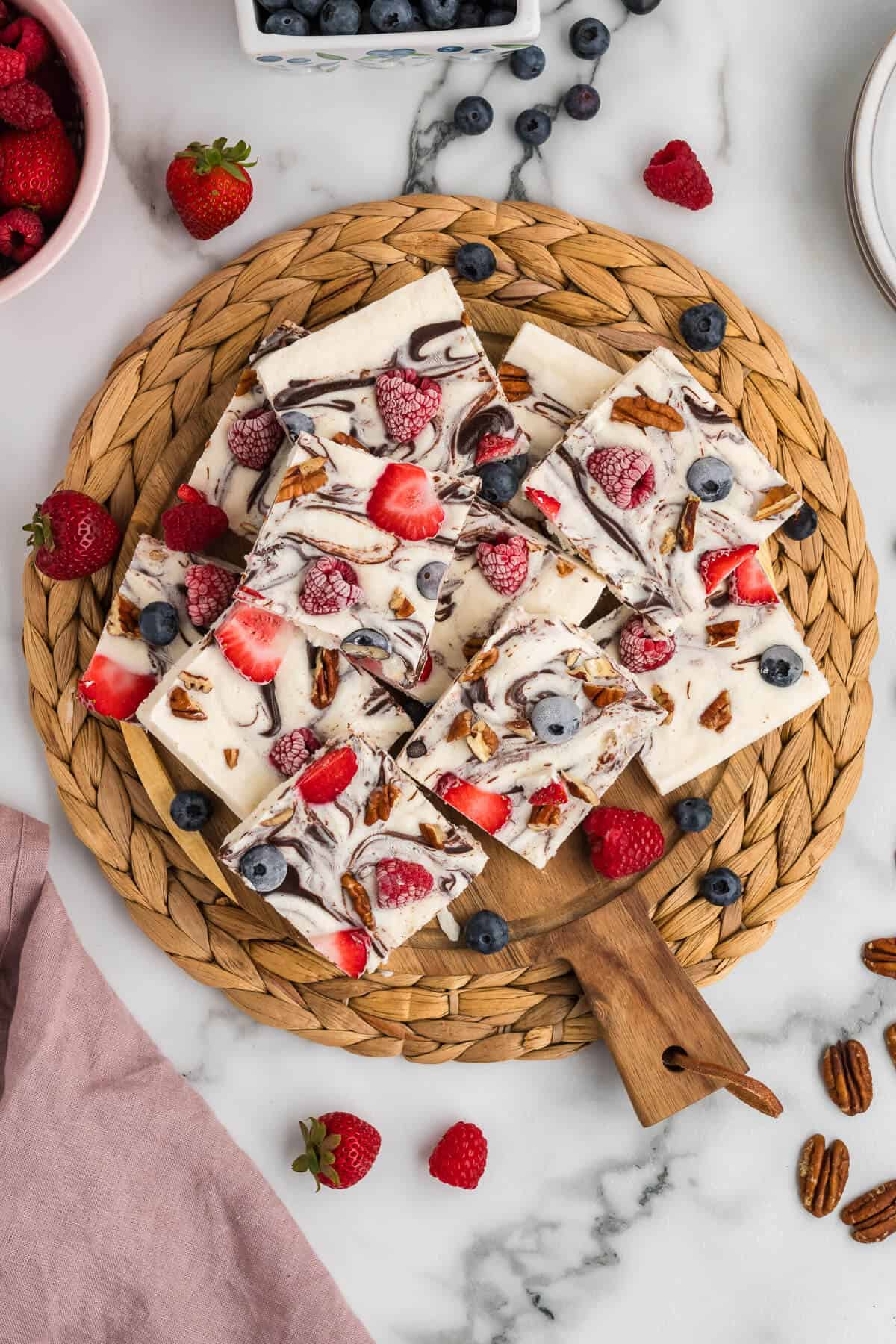 Frozen Yogurt Bark with berries and pecans on a round wooden board, garnished with some fresh berries on top and scattered around.