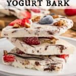 Three squares of Frozen Yogurt Bark stacked on a plate. Remaining Yogurt Bark is in the back ground. The frozen bark has berries, pecans and chocolate in it. Text overlay reads: Frozen Yogurt Bark