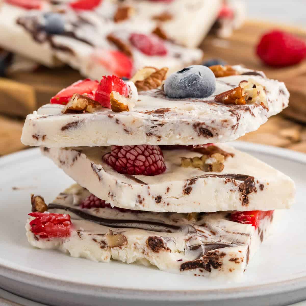 Three squares of Frozen Yogurt Bark stacked on a plate. Remaining Yogurt Bark is in the back ground. The frozen bark has berries, pecans and chocolate in it.