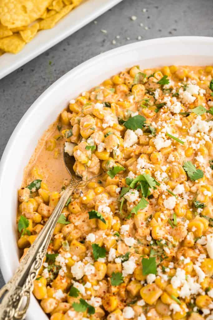 Elote dip in a round serving dish, garnished with crumbled cotija cheese and fresh cilantro. A spoon lies towards the side of the dish scooped with the corn dip.