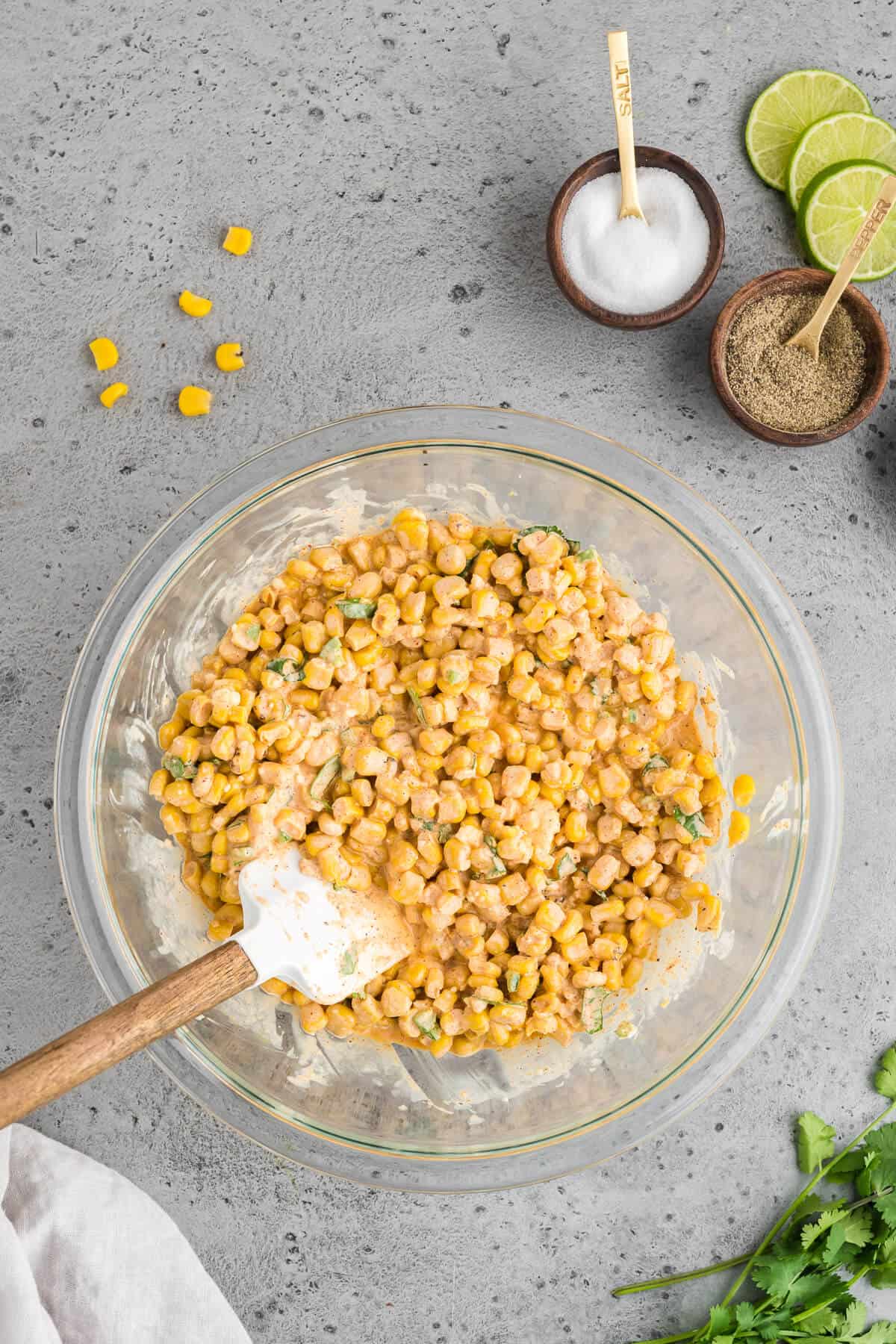 Elote dip being stirred in a mixing bowl. Salt, pepper, and lime slices are off to the side.