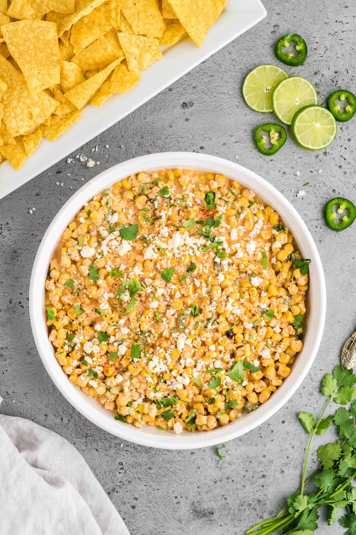 Elote dip in a round serving dish surrounded with chips, some lime slices and jalapeno slices, and some fresh cilantro.