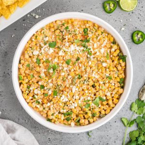 Elote dip in a round serving dish surrounded with chips, some lime slices and jalapeno slices, and some fresh cilantro.