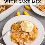 Peach dump cake on a white plate topped with vanilla ice cream and sliced almonds. The pan of cake is in the back ground, a few fresh peach slices scattered around. Text overlay reads: Peach Cobler with Cake Mix