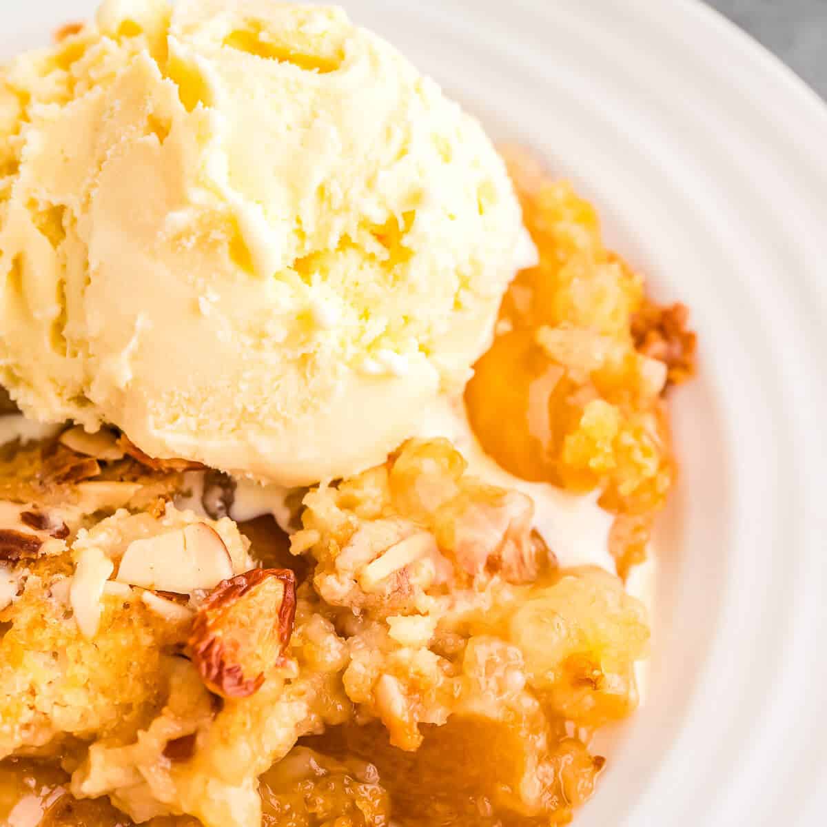 Close up view of Peach Dump cake on a white plate topped with a scoop of vanilla ice cream that's just beginning to melt.