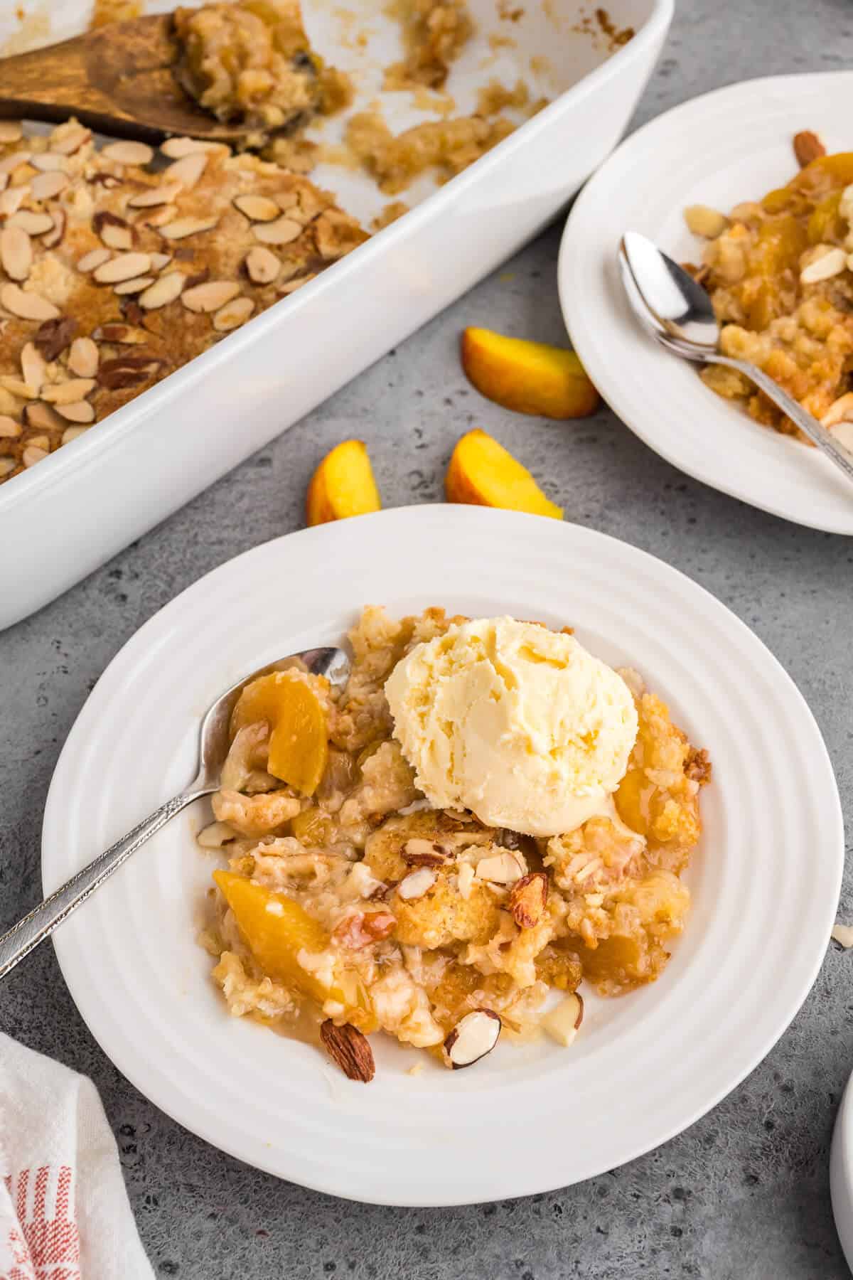 Two plates of peach dump cake with sliced almonds on white plates with a scoop of vanilla ice cream on top. The pan of cobbler is in the back ground.