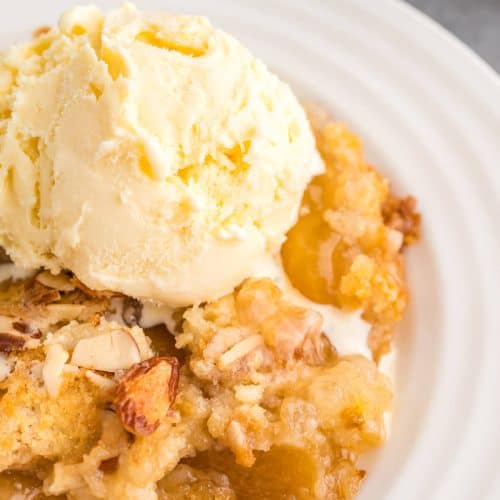 Close up view of Peach Dump cake on a white plate topped with a scoop of vanilla ice cream that's just beginning to melt.