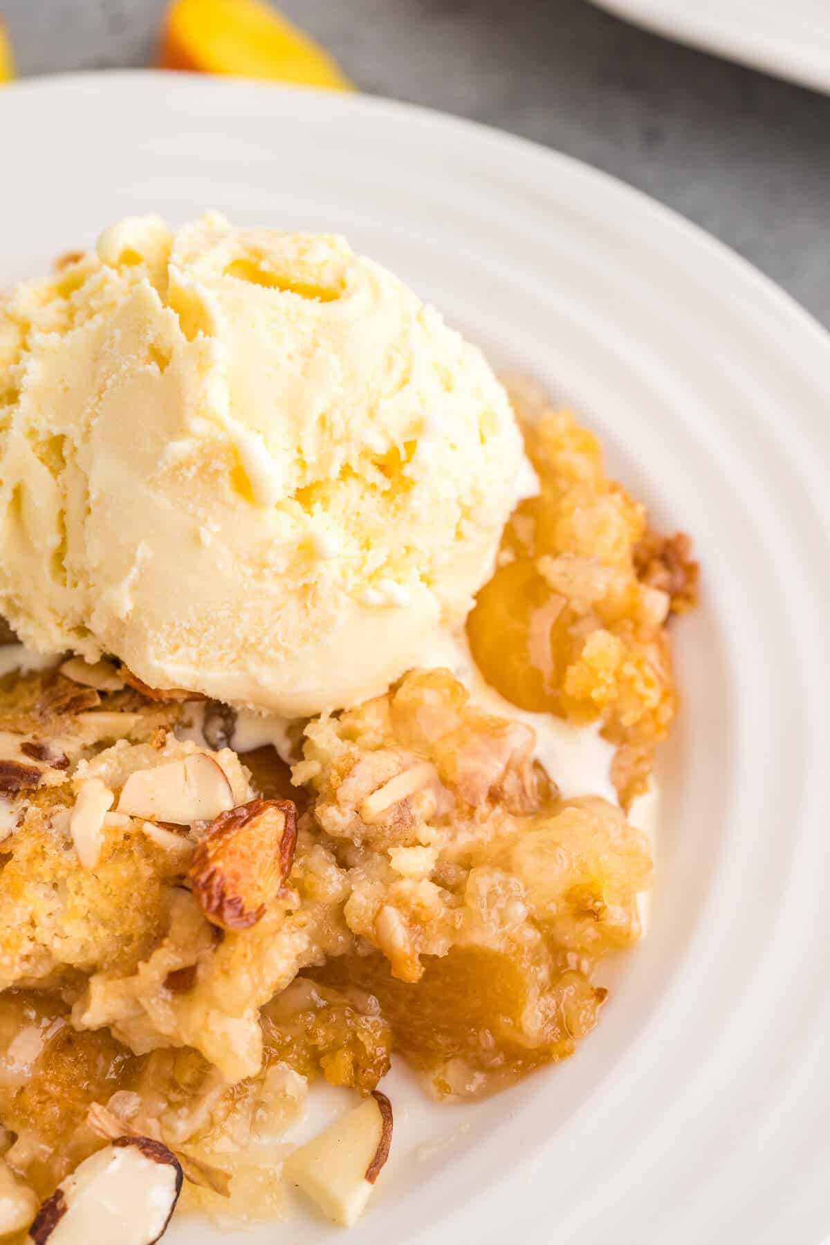 Close up view of Peach Dump cake on a white plate topped with a scoop of vanilla ice cream that's just beginning to melt.