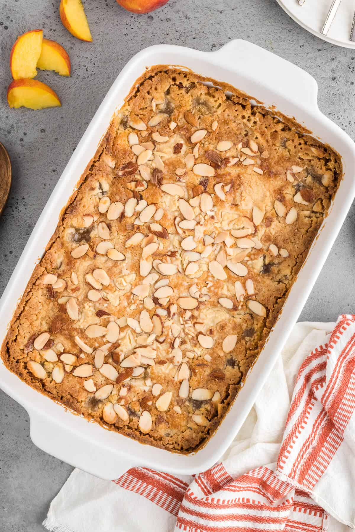 Peach Dump Cake in a white casserole dish. The top of the cake is lightly browned and sprinkled with sliced almonds. There are a few slices of fresh peaches around one side of the dish.