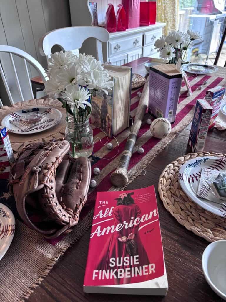 Baseball themed table scape. The book "The All American, baseball bat, glove and baseballs make a centerpiece. There is also chocolate candy wrapped to look like baseballs scattered about. An American Flag Burlap runner is the base. There is also a boquet of daisies, which ties into the book, some classic novels, baseball plates and napkins.