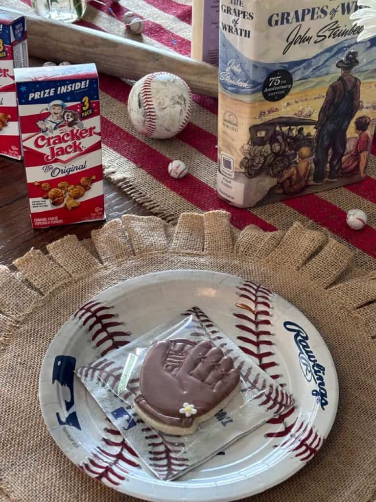 Baseball place setting of burlap placemat, baseball plate, napkin, and decorated cookie of a baseball glove. Cracker Jacks and a used baseball in the background.