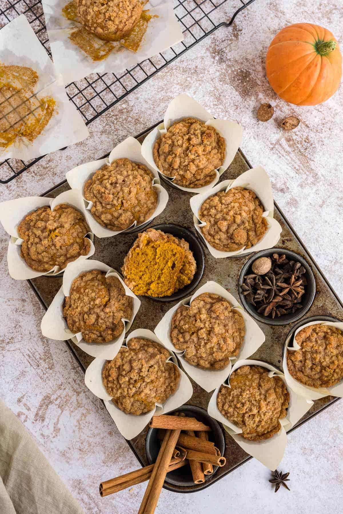 Fresh baked Pumpkin Streusel Muffins in the muffin tin. One well shows a half of muffin to see the inside. another well contains star anise and whole nutmeg, and another well contains cinnamon sticks.