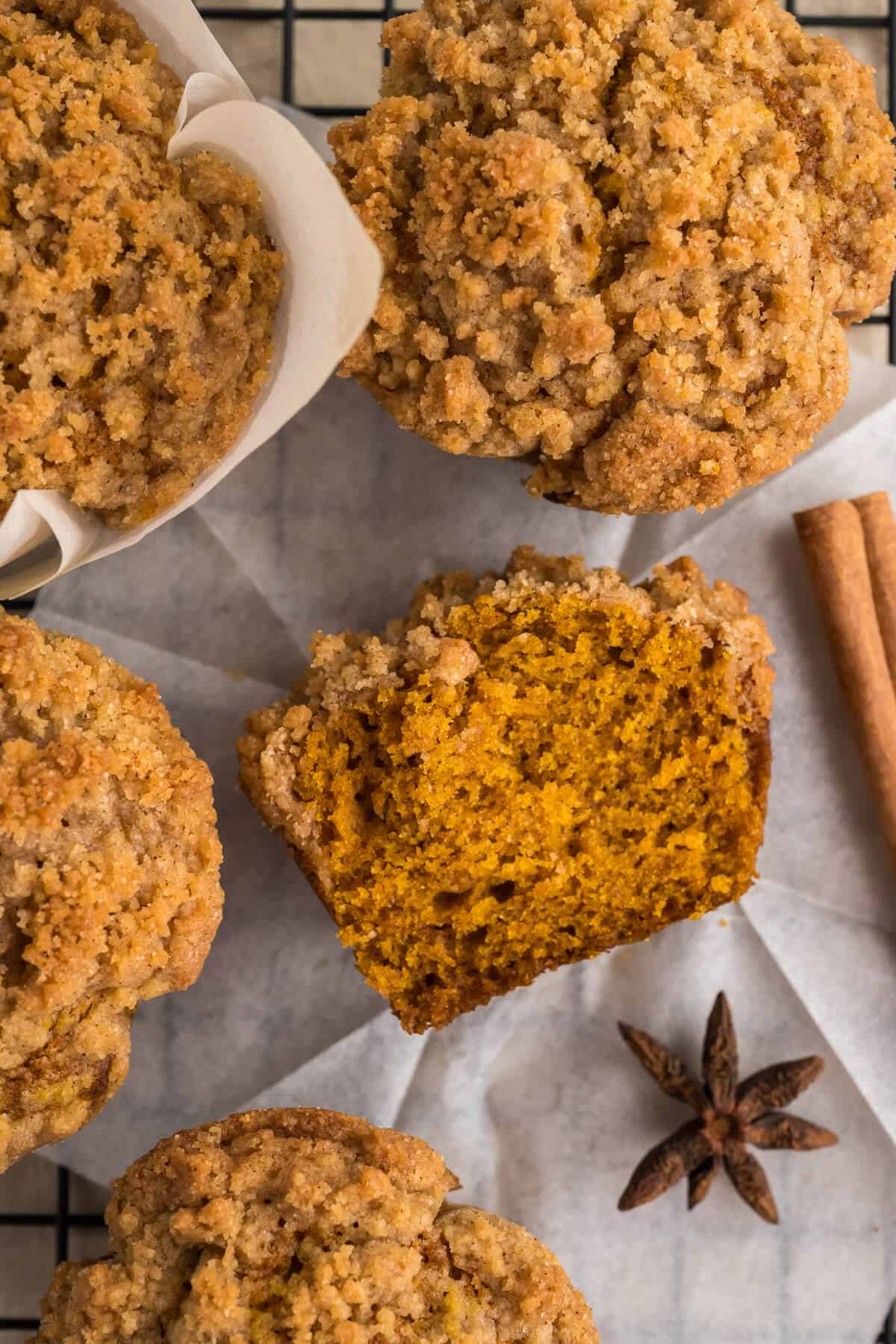 Overhead view of Pumpkin streusel muffins on a baking rack lined with parchment paper. One muffin is cut in half laying on its side. Cinnamon stick and star anise are off to the side as a garnish.