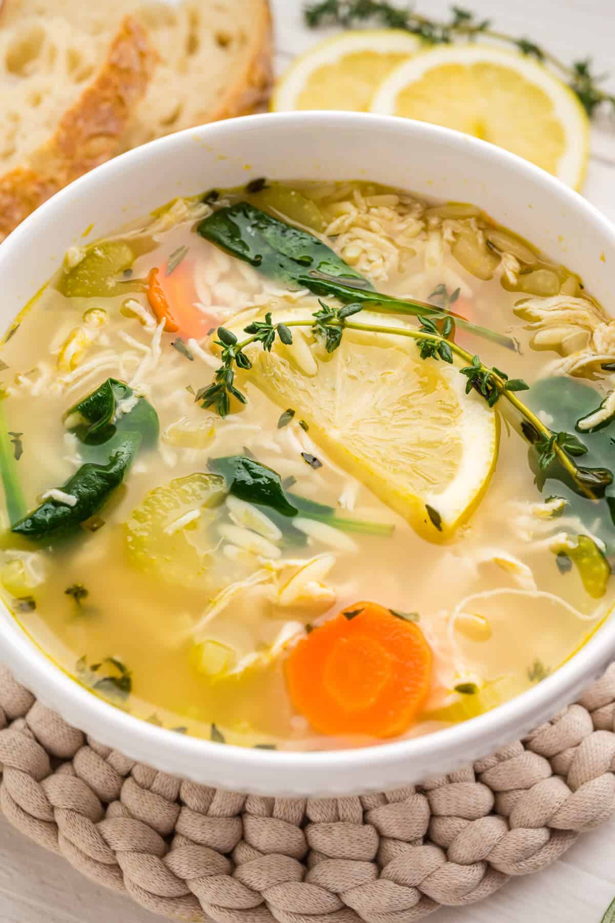 Close up image of a bowl of Lemon Chicken Orzo Soup with spinach, garnished with a lemon slice and fresh sprig of thyme. Crusty bread is served along side it.