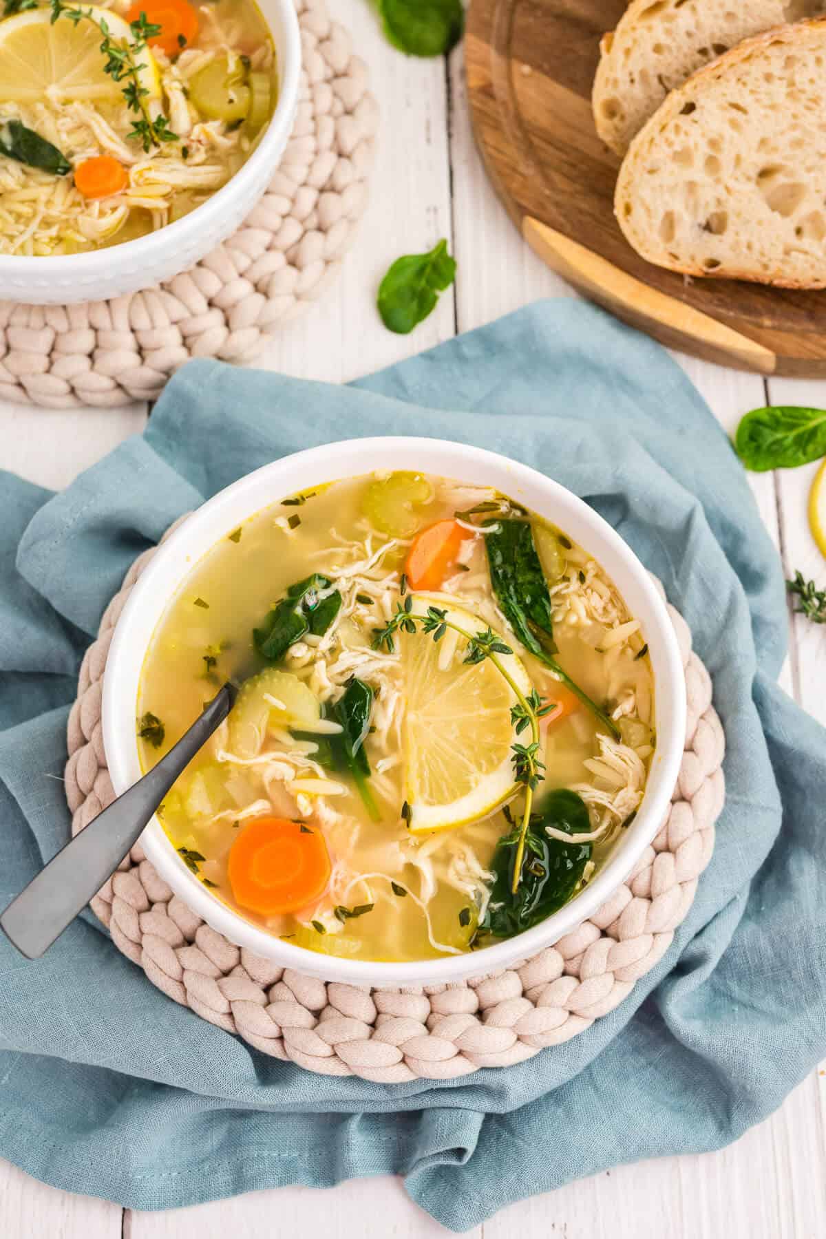 Two white bowls of Lemon Orzo Soup, garnished with a sprig of thyme and a lemon slice. A spoon is resting in the bowl. A few slices of sourdough bread are in the background.