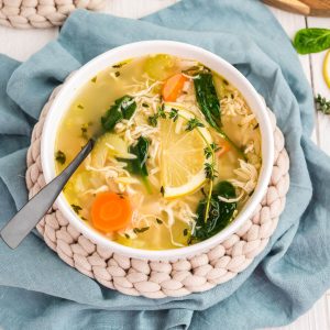 A bowl of Lemon Orzo Soup, garnished with a sprig of thyme and a lemon slice. A spoon is resting in the bowl. The bowl sits on top of a woven trivet, which is on top of a light blue linen.