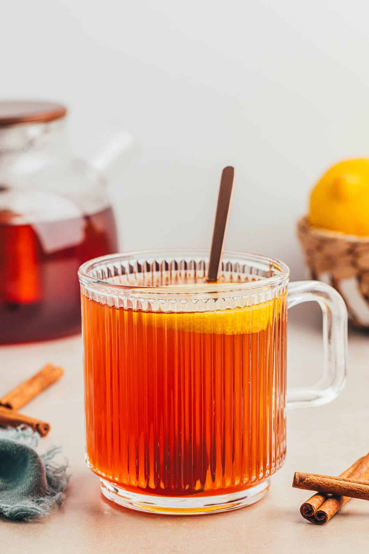 A clear ribbed mug of a non alcoholic hot toddy with a slice of lemon floating on top and a spoon is in the mug. A small pot of tea sits in the background.