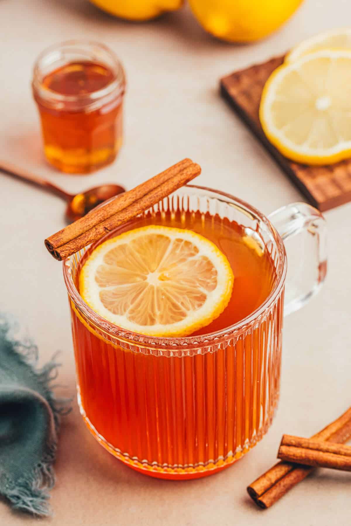 A clear ribbed mug of a non alcoholic hot toddy with a slice of lemon floating on top and a cinnamon stick resting on the edge of the mug. Another small jar of honey and lemon slices sit in the background.