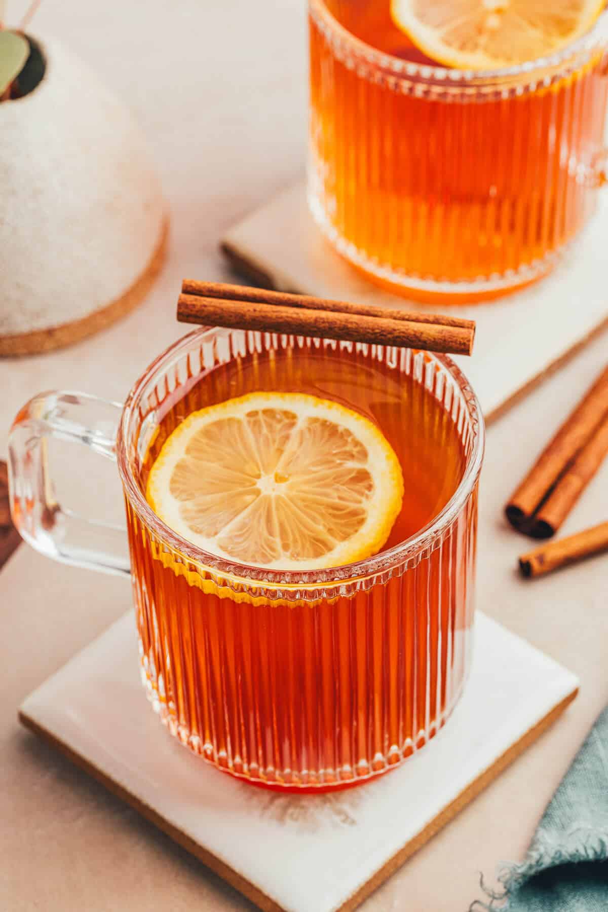 A clear ribbed mug of a non alcoholic hot toddy with a slice of lemon floating on top and a cinnamon stick resting on the edge of the mug. Another mug of tea sits in the background.