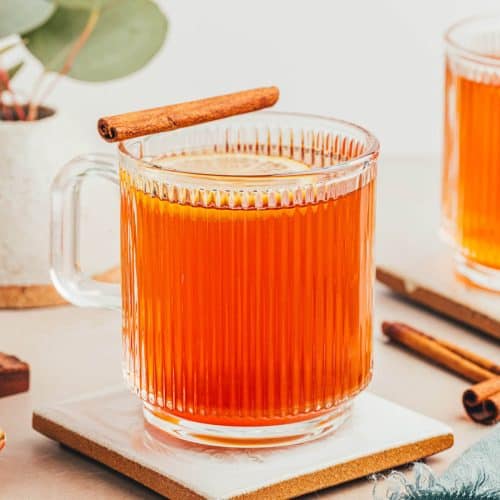 2 mugs of a hot toddy made without alcohol. The mugs are clear and ribbed giving a beautiful view of the warm drink which is amber in color. Lemon slices float on top and cinnamon sticks balance on the edge.