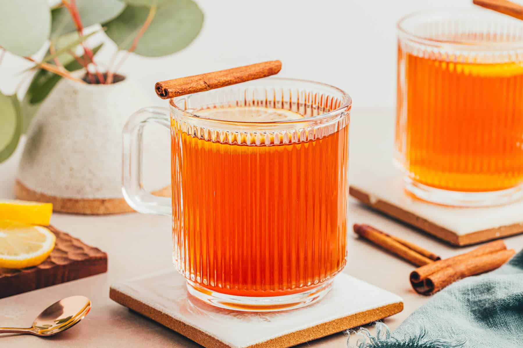 2 mugs of a hot toddy made without alcohol. The mugs are clear and ribbed giving a beautiful view of the warm drink which is amber in color. Lemon slices float on top and cinnamon sticks balance on the edge.