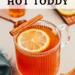 A clear ribbed mug of a non alcoholic hot toddy with a slice of lemon floating on top and a cinnamon stick resting on the edge of the mug. Another small jar of honey and lemon slices sit in the background. Text overlay reads: Non alcoholic hot toddy